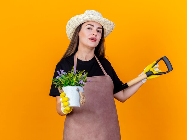 Menina linda jardineira satisfeita vestindo uniforme e chapéu de jardinagem com luvas, colocando a pá no ombro e segurando uma flor em um vaso de flores isolado em um fundo laranja