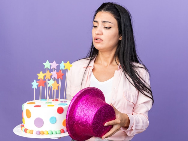 Menina linda jovem insatisfeita segurando um bolo segurando um chapéu e olhando para o bolo na mão