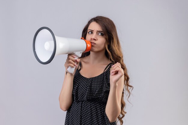 Menina linda num vestido de bolinhas descontente gritando para o megafone com expressão de raiva em pé