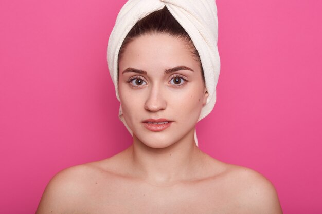 Foto grátis menina linda, vestindo uma toalha branca na cabeça, posando com os ombros à mostra, isolados no estúdio rosa, senhora atraente olhando diretamente para a câmera, tendo a pele limpa, perfeita. conceito de cuidados com a pele.