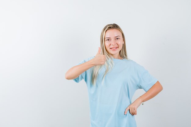 Menina loira aparecendo o polegar, segurando a mão na cintura em uma camiseta azul e parecendo confiante, vista frontal.