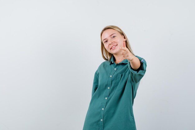 Menina loira apontando para a câmera com o dedo indicador em uma blusa verde e parecendo feliz