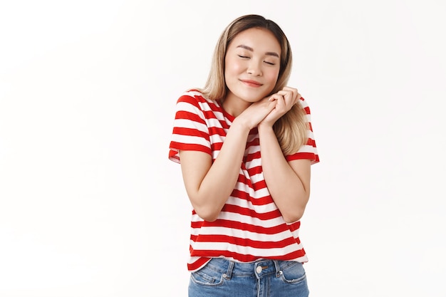 Foto grátis menina loira asiática fofa e sonhadora fechar os olhos sorrindo feliz romanticamente imaginando férias de verão perfeitas apertando as mãos, animado, lembra-se do bom momento