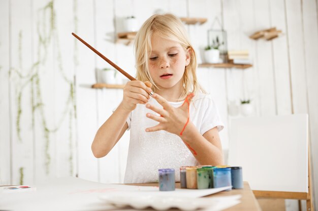 Menina loira bonita e bonita em camiseta branca, alegremente, pintando a palma da mão com pincel, em pé atrás da mesa com jarra de água, pincéis e tinta sobre ele.