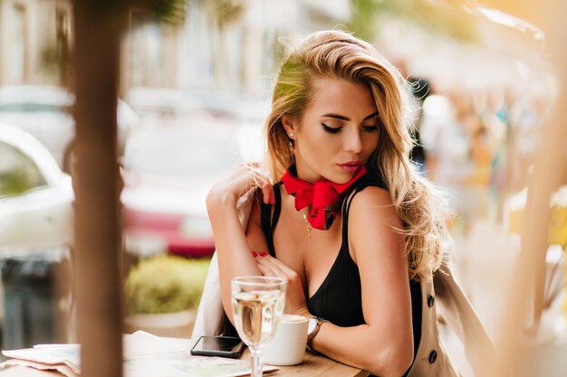 Menina loira bronzeada relaxando em um café ao ar livre com uma xícara de chá e olhando para baixo. Mulher atraente de cabelos louros em um vestido preto, relaxando em um restaurante localização com smartphone e jornal.