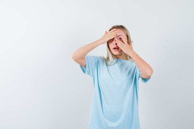 Menina loira cobrindo os olhos com as mãos, olhando por entre os dedos em uma camiseta azul e bonita, vista frontal.