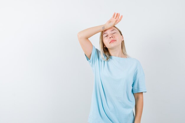 Menina loira colocando a mão na testa, tendo dor de cabeça na camiseta azul e parecendo exausta. vista frontal.