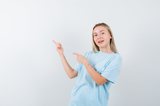 Menina loira com camiseta azul apontando para a esquerda com o dedo indicador e parecendo feliz, vista frontal.