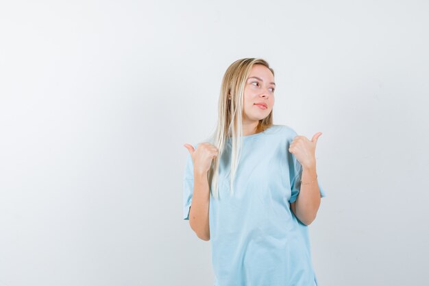 Menina loira com camiseta azul apontando para o lado com os polegares e parecendo feliz, vista frontal.