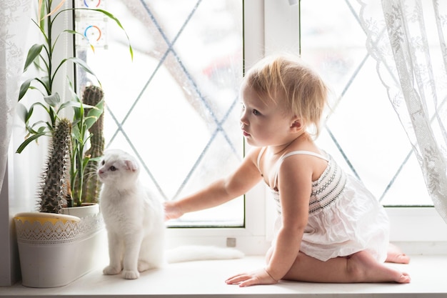 Foto grátis menina loira criança brincando com gato branco na janela conceito de estilo de vida interior real da infância e animais