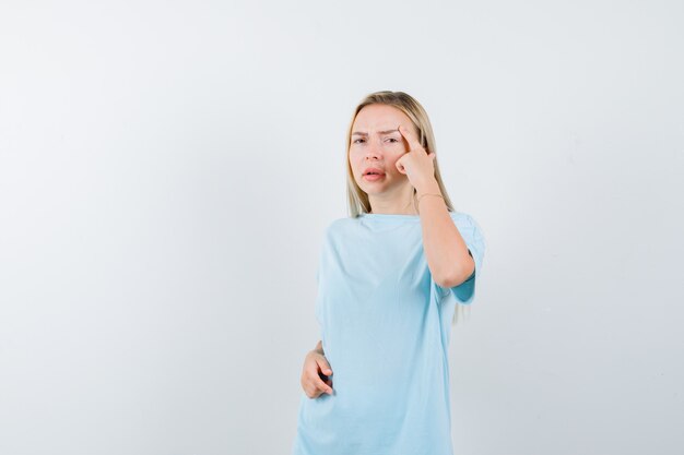 Menina loira de camiseta azul, colocando o dedo indicador na têmpora e olhando pensativa, vista frontal.