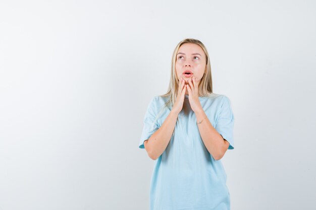 Menina loira de camiseta azul de mãos dadas no queixo, olhando para cima e parecendo surpresa, vista frontal.