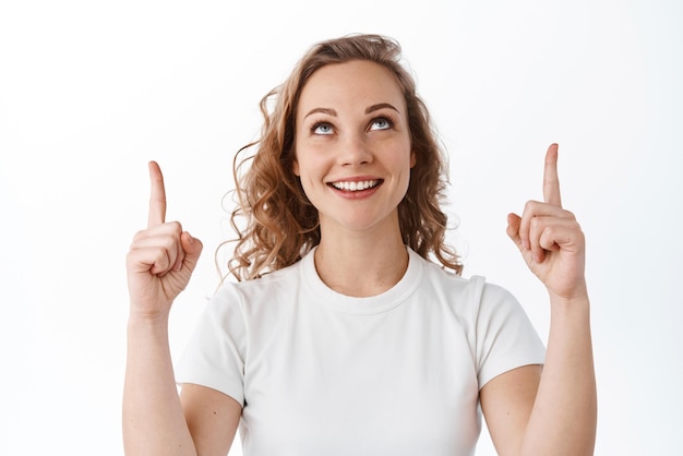 Menina loira elegante com penteado encaracolado sorrindo satisfeito apontando e olhando para o fundo branco do anúncio