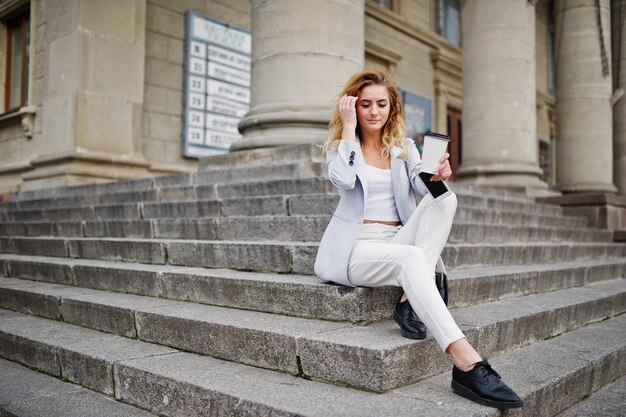 Menina loira elegante e encaracolada veste branco com uma xícara de café posando nas escadas ao ar livre