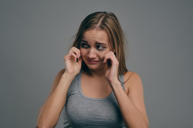 Menina loira em pânico com cabelos desgrenhados e hematomas está tocando seu rosto em camiseta cinza posando contra o fundo cinza do estúdio abuso de violência doméstica depressão desespero espaço de cópia de closeup