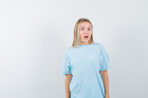 Menina loira em pé e posando para a câmera em t-shirt azul e bonita, vista frontal.
