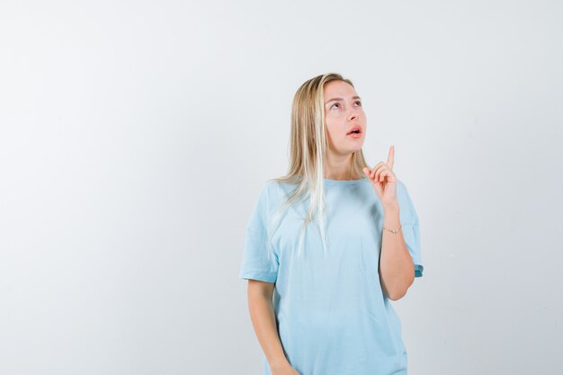 Menina loira em uma camiseta azul apontando para cima com o dedo indicador, olhando para cima e olhando pensativa, vista frontal.