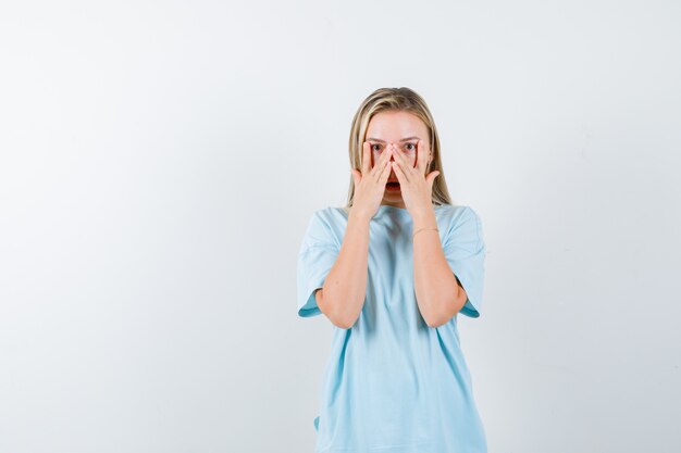 Menina loira em uma camiseta azul mostrando sinais de v nos olhos, olhando por entre os dedos e parecendo surpresa, vista frontal.