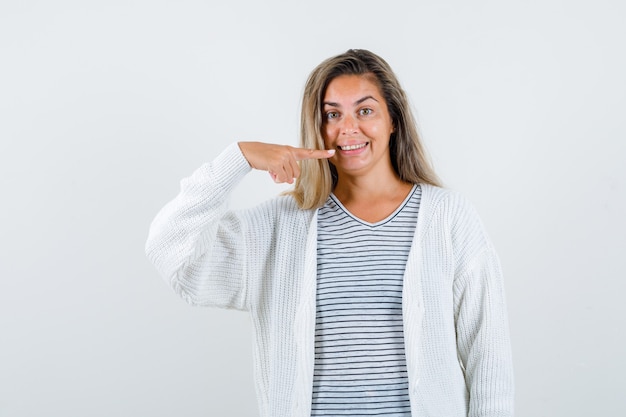 Menina loira em uma camiseta listrada, casaco de lã branco e calças jeans apontando para os dentes e parecendo feliz, vista frontal.