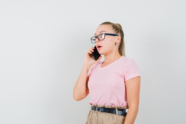 Menina loira falando no celular em t-shirt, calça e olhando pensativa, vista frontal.