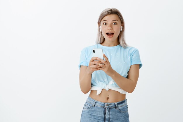 Menina loira impressionada e satisfeita posando no estúdio com seu telefone e fones de ouvido