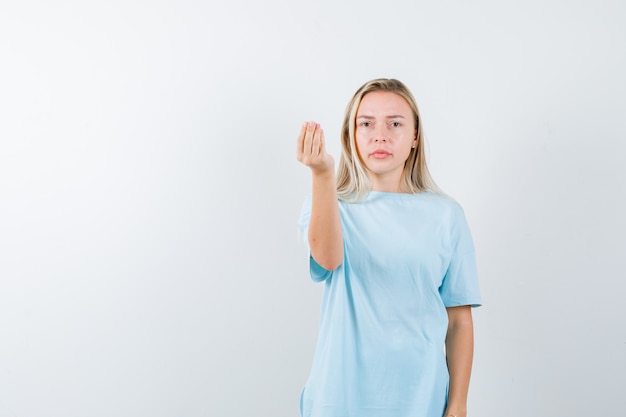 Menina loira mostrando gesto italiano em t-shirt azul e olhando sério, vista frontal.