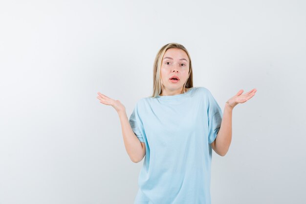 Menina loira mostrando um gesto impotente em t-shirt azul e parecendo confusa. vista frontal.