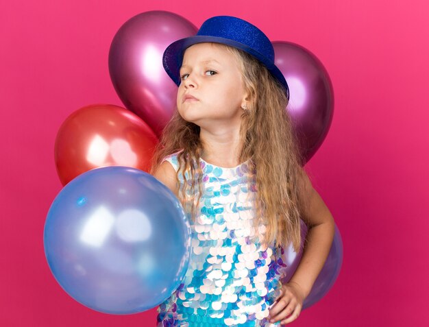 menina loira ofendida com chapéu de festa azul em pé com balões de hélio isolados na parede rosa com espaço de cópia