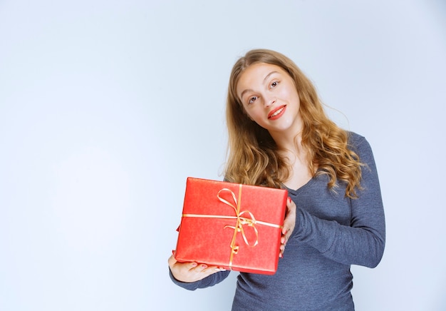 Menina loira oferecendo uma caixa de presente vermelha para alguém.