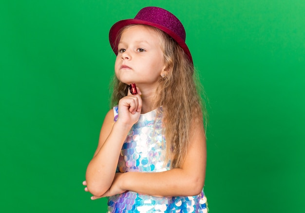 Foto grátis menina loira pensativa com chapéu de festa roxo colocando a mão no queixo segurando o apito isolado na parede verde com espaço de cópia