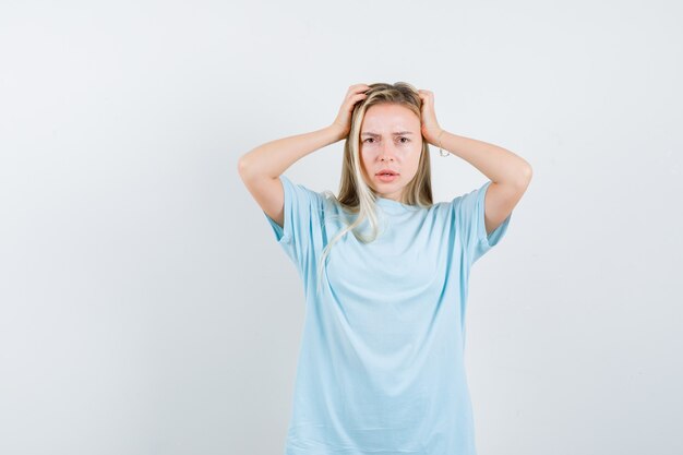 Menina loira segurando a cabeça com as mãos na camiseta azul e olhando séria, vista frontal.