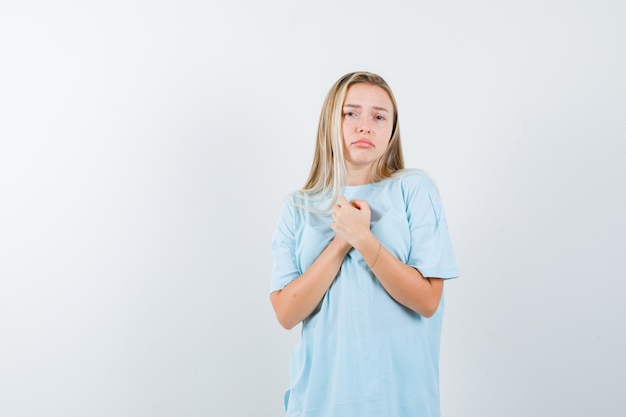 Menina loira segurando as mãos sobre o peito em t-shirt azul e olhando preocupada, vista frontal.