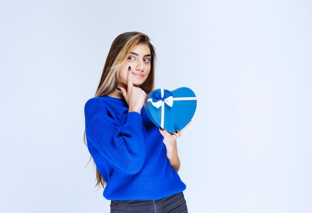 Menina loira segurando uma caixa de presente de forma de coração azul e pensando.