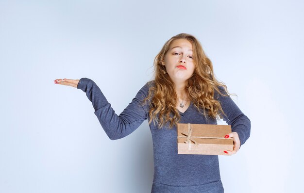 Menina loira segurando uma caixa de presente de papelão e parece confusa e pensativa.