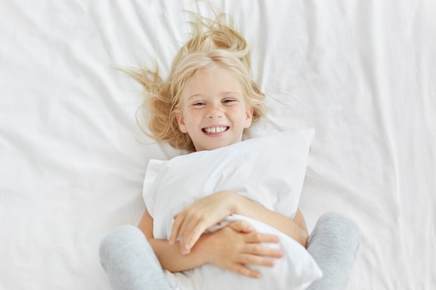 Menina loira sorridente, abraçando o travesseiro branco enquanto estiver no jardim de infância, tendo bom humor ao ver alguém e deitado na cama branca. Adorável criança do sexo feminino ter hora de dormir. Conceito de descanso