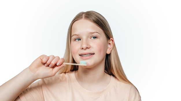 Menina loira sorridente com aparelho ortodôntico segurando uma escova de dentes de bambu ecológica
