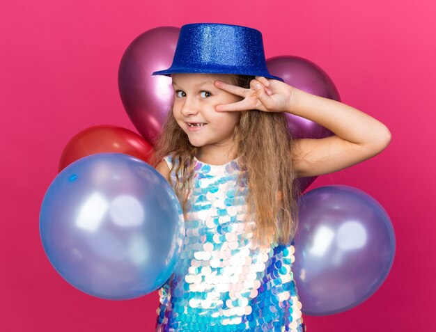 menina loira sorridente com chapéu de festa azul segurando balões de hélio e gesticulando o sinal da vitória isolada na parede rosa com espaço de cópia