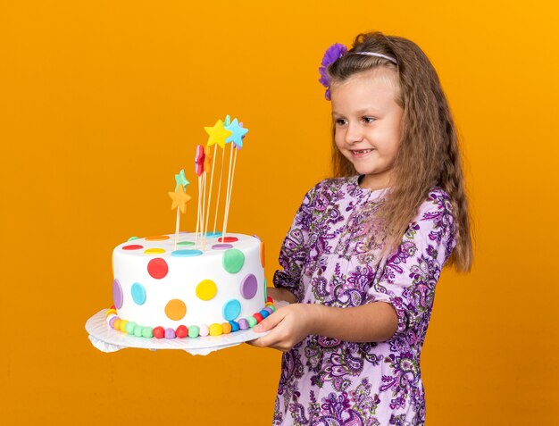 menina loira sorridente segurando e olhando para um bolo de aniversário isolado na parede laranja com espaço de cópia