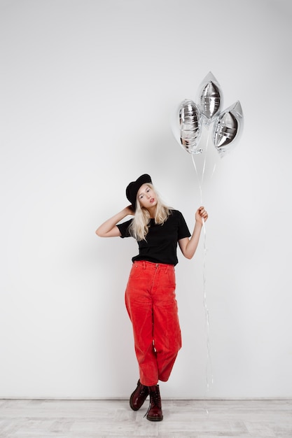 Menina loura bonita nova que guarda baloons de prata sobre a parede branca.