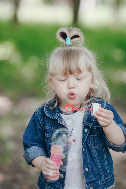 Menina loura pequena soprando por uma bolhas pomposos e fazendo