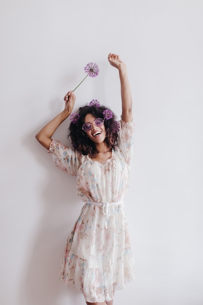 Menina magro com cabelo preto ondulado dançando em casa e sorrindo. Mulher jovem africana relaxante em um vestido romântico, posando com flores.