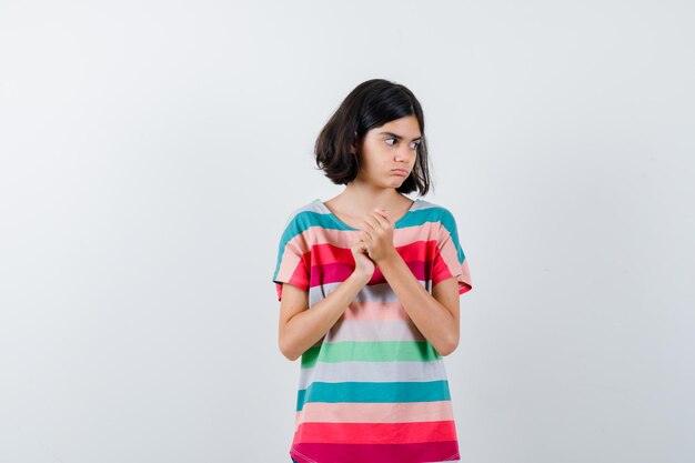 Menina, mantendo as mãos cruzadas na camiseta e olhando pensativa, vista frontal.