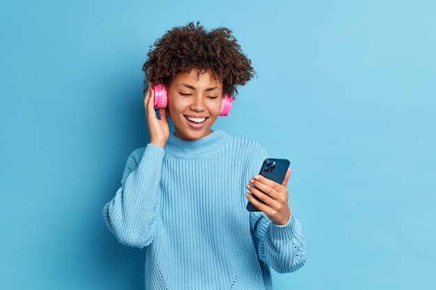Menina milenar positiva entretida com cabelo encaracolado segurando smartphone faz videochamada e usa fones de ouvido estéreo para ouvir suas músicas favoritas vestida com um macacão quente