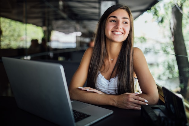 Menina modelo sorridente e ensolarada trabalhando em seu laptop no terraço do café daytilme