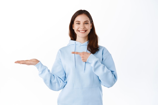 Menina morena alegre apontando para a palma da mão segurando copyspace, mostrando o item em exibição em sua mão, em pé contra uma parede branca