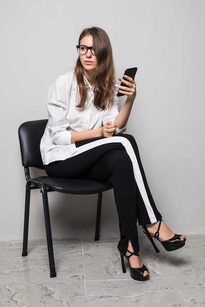 Menina morena alta de óculos, vestida com uma camiseta branca e calça preta, sentada com o telefone na cadeira do escritório na frente do fundo branco