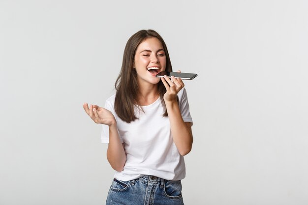 Menina morena atraente amigável falando no alto-falante, gravar mensagem de voz no smartphone, branco.