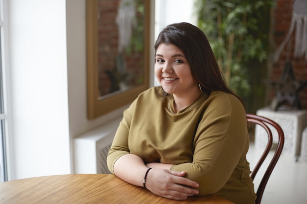 Foto grátis menina morena atraente positiva com corpo curvilíneo largy, sentado à mesa do café sozinha, esperando seu almoço. foto de uma jovem linda e gorda posando em um interior aconchegante e elegante, sorrindo feliz