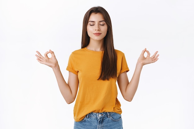 Menina morena bonita calma e relaxada para liberar o estresse, fechar os olhos e sorrir pacificamente, meditando com as mãos abertas para os lados em mudra, zen, pose de lótus em pé, prática de ioga, fundo branco