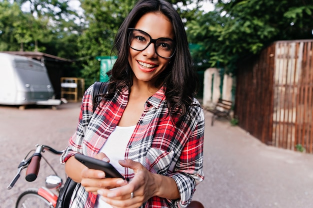 Menina morena curiosa com o telefone na mão olhando. Foto ao ar livre de uma deslumbrante senhora latina ao lado da bicicleta.